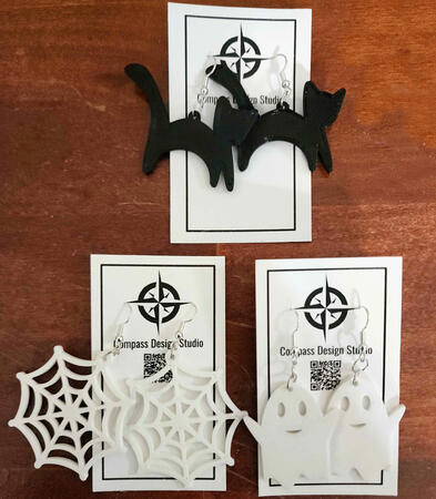 Three pairs of earrings with silver ear hooks - one of a black cat silhouette, one of a smiling ghost, and one of a spiderweb
